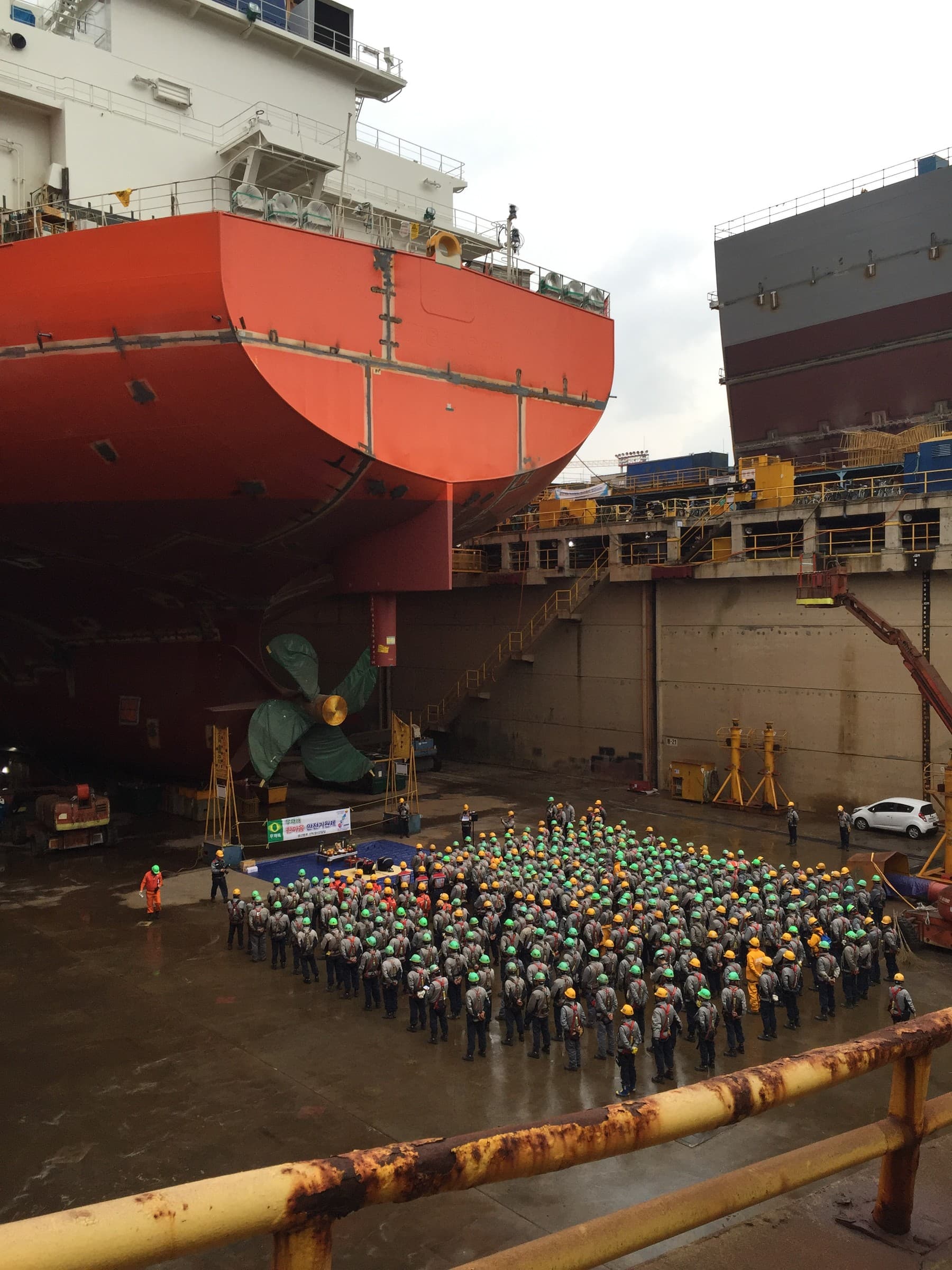 Shipyard workers gathered in a dry dock beneath a large ship's stern